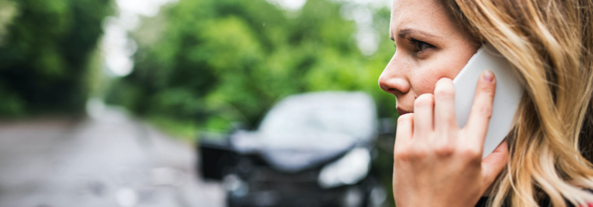 Close up of woman holding a phone to her ear. In the background is a black sedan with a smashed front-end.