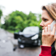 Close up of woman holding a phone to her ear. In the background is a black sedan with a smashed front-end.