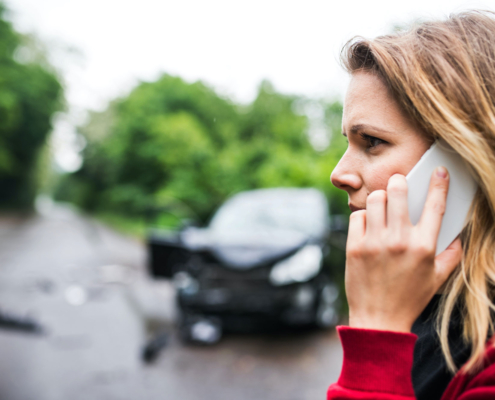 Close up of woman holding a phone to her ear. In the background is a black sedan with a smashed front-end.