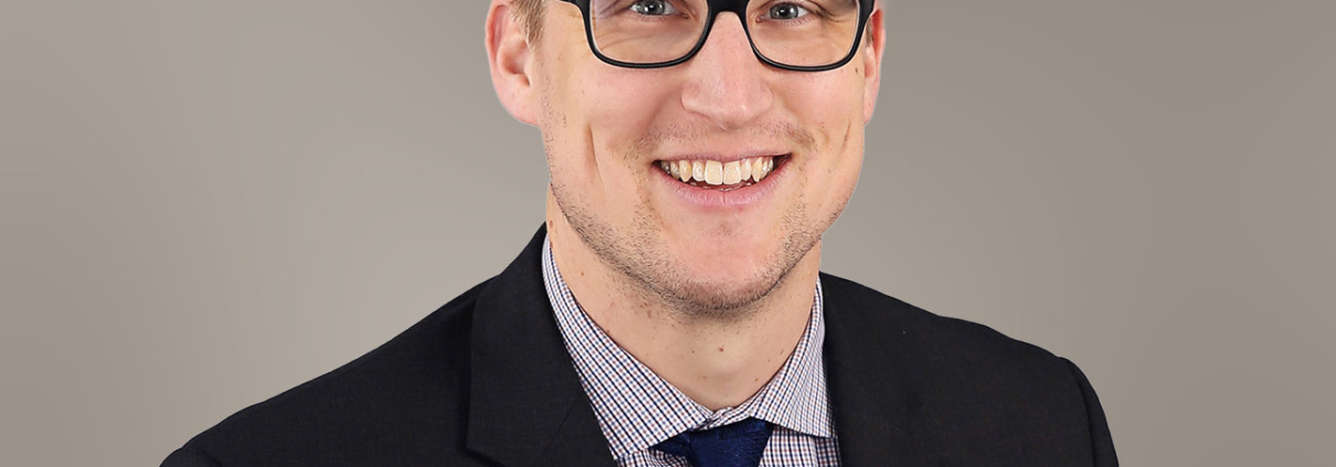 Headshot of Corey Olson in the foreground. Corey is wearing a black suit coat and collared shirt with tie. The background color is taupe.