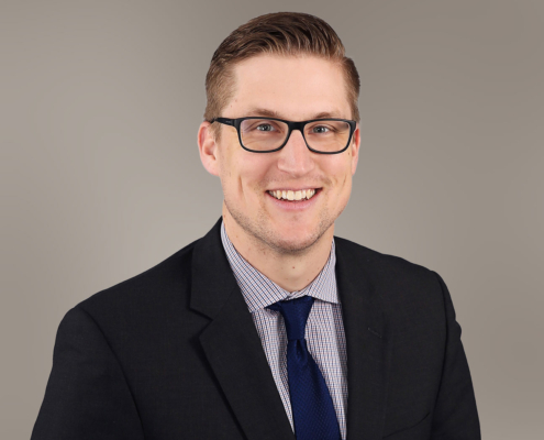 Headshot of Corey Olson in the foreground. Corey is wearing a black suit coat and collared shirt with tie. The background color is taupe.