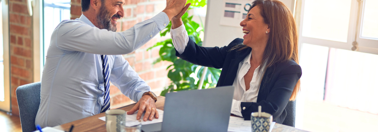 Man and woman in a business setting. They are sitting next to each other at a table, and a laptop is open in front of them. They are high-fiving each other.