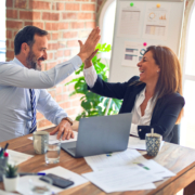 Man and woman in a business setting. They are sitting next to each other at a table, and a laptop is open in front of them. They are high-fiving each other.
