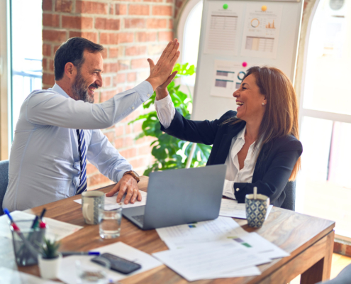 Man and woman in a business setting. They are sitting next to each other at a table, and a laptop is open in front of them. They are high-fiving each other.