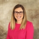 Headshot of Carrie Wingenbach. Carrie is wearing a long-sleeved coral pink shirt. The photo background is tan.