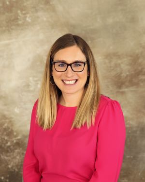 Headshot of Carrie Wingenbach. Carrie is wearing a long-sleeved coral pink shirt. The photo background is tan.