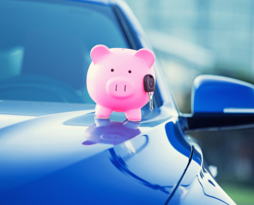 A pink piggy bank holding a car key sits on the hood of a blue car