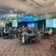 Green and blue stage lighting against black background features the HR Collaborative logo. On the stage is a podium with a white front and showing the HR Collaborative's green and blue logo. The room is filled with round tables covered in a black tablecloth, and six people are seated at each table.