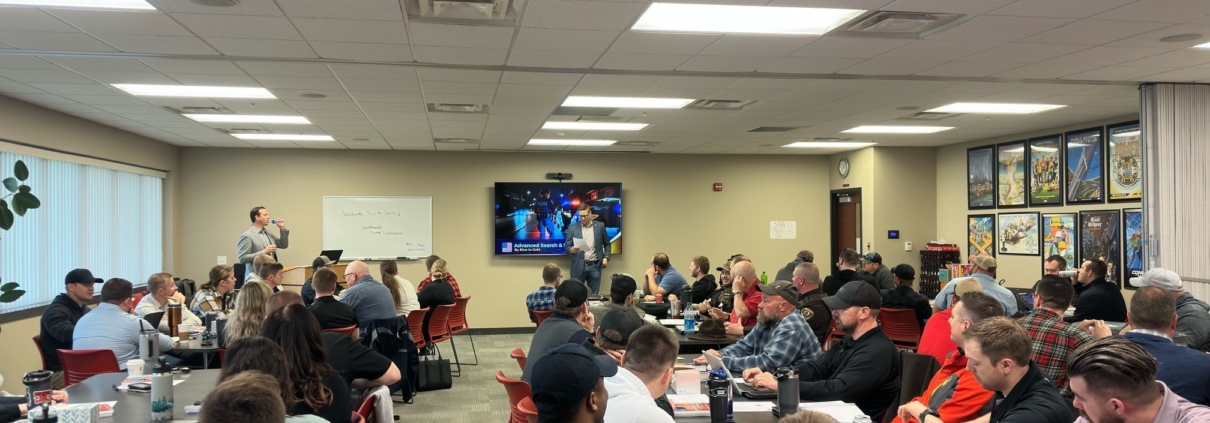 Director of Member Services Corey Olson stands at the front of a large classroom in filled with people sitting in chars at tables placed throughout the room.