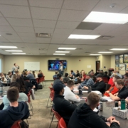 Director of Member Services Corey Olson stands at the front of a large classroom in filled with people sitting in chars at tables placed throughout the room.
