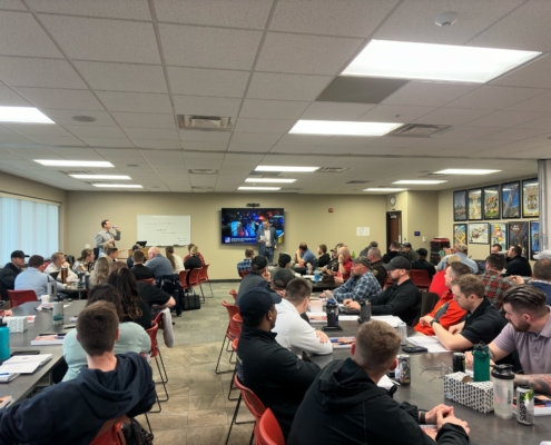 Director of Member Services Corey Olson stands at the front of a large classroom in filled with people sitting in chars at tables placed throughout the room.