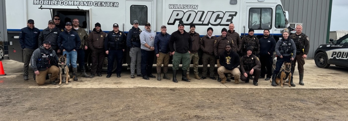 A group of 20 law enforcement officers, including two K9 officers, pose for a photo in front of the Williston Police Department's white Mobile Command Center trailer.