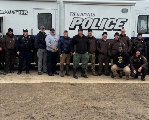 A group of 20 law enforcement officers, including two K9 officers, pose for a photo in front of the Williston Police Department's white Mobile Command Center trailer.