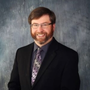 Jordan Wahl wears a black suit coat with purple tie and black wire-rimmed glasses while smiling for his professional headshot. The background is a gray blue color.