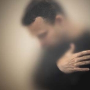 Blurred photo of a man wearing a black long-sleeved shirt while looking down and holding his arm over his chest.