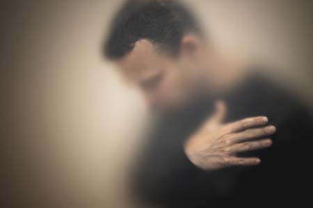 Blurred photo of a man wearing a black long-sleeved shirt while looking down and holding his arm over his chest.